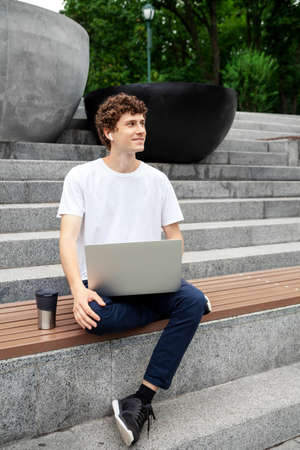 European young male in wireless earphones wearing white t-shirt sitting in a city park and looking in smartphone. Freelance work concept outdoors. Empty copy space.elance work concept outdoors.の写真素材