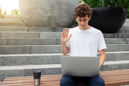 European young male in wireless earphones wearing white t-shirt sitting in a city park and looking in smartphone. Freelance work concept outdoors. Empty copy space.elance work concept outdoors.の写真素材