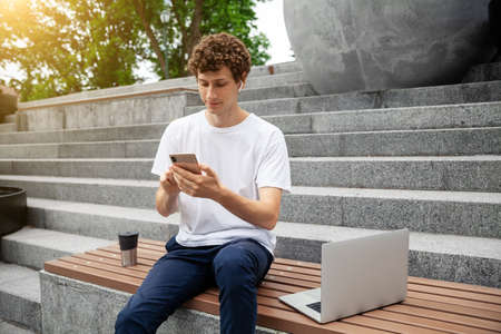 European young male in wireless earphones wearing white t-shirt sitting in a city park and looking in smartphone. Freelance work concept outdoors. Empty copy space.elance work concept outdoors.の写真素材