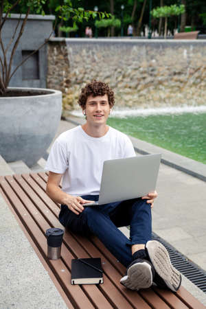 European young male in wireless earphones wearing white t-shirt sitting in a city park and looking in smartphone. Freelance work concept outdoors. Empty copy space.elance work concept outdoors.の写真素材