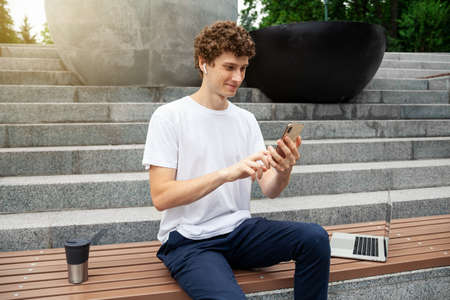 European young male in wireless earphones wearing white t-shirt sitting in a city park and looking in smartphone. Freelance work concept outdoors. Empty copy space.elance work concept outdoors.の写真素材