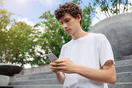 European young male in wireless earphones wearing white t-shirt sitting in a city park and looking in smartphone. Freelance work concept outdoors. Empty copy space.elance work concept outdoors.の写真素材
