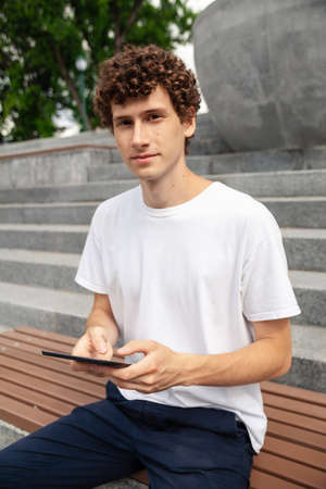 European young male in wireless earphones wearing white t-shirt sitting in a city park and looking in smartphone. Freelance work concept outdoors. Empty copy space.elance work concept outdoors.の写真素材