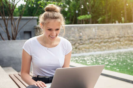 Dreamy and stylish European young student girl in wireless earphones wearing white t-shirt, writing to notebook at summer day.の写真素材