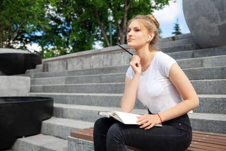 Dreamy and stylish European young student girl in wireless earphones wearing white t-shirt, writing to notebook at summer day.の写真素材