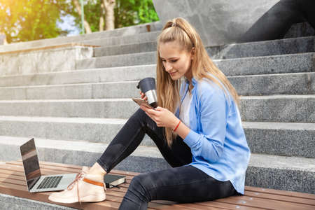 Dreamy and stylish European young student girl in wireless earphones wearing white t-shirt, writing to notebook at summer day.の写真素材