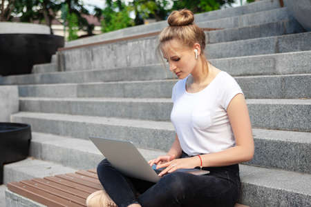 Dreamy and stylish European young student girl in wireless earphones wearing white t-shirt, writing to notebook at summer day.の写真素材