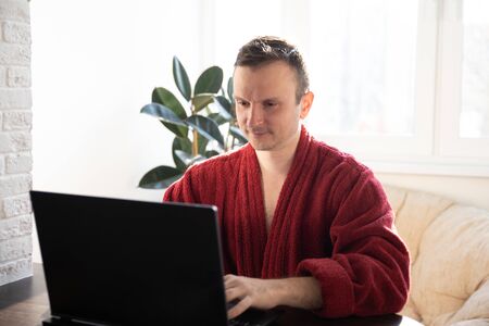 A young man smiles as he reads on a laptop screen while relaxing on a comfortable sofa at home in dressing gownsの写真素材