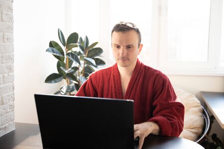 A young man smiles as he reads on a laptop screen while relaxing on a comfortable sofa at home in dressing gownsの写真素材
