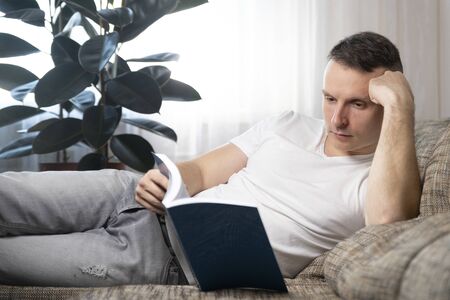 Leisure, education and the main concept-a smiling guy reads a book and sits at home on the couch. Rest, relaxation.の写真素材