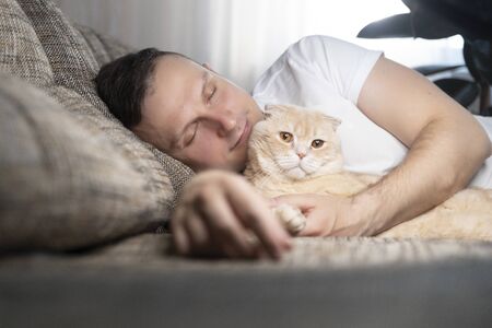 A young man falls asleep on a sofa with a book. Next to him is his beautiful ginger cat. The concept of rest and relaxationの写真素材