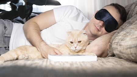 A young man falls asleep on a sofa with a book. Next to him is his beautiful ginger cat. The concept of rest and relaxationの写真素材