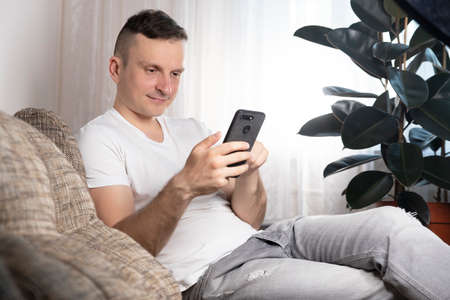 A young smiling man checks social networks on a smartphone at home while sitting on the sofa. Stylish boy in casual clothes, copy of spaceの写真素材