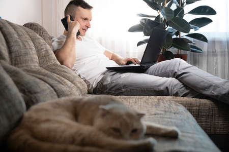 The cat lies on the couch, a man works on a laptop. A young quarantined guy is working at home, with a red cat lying nearby. Remote work, work from home.の写真素材