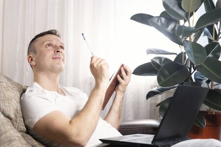 A focused European male freelancer takes notes studies working with a laptop, a young professional writes an essay in a notebook prepares for a test exam and sits at home on the couch. Remote workの写真素材
