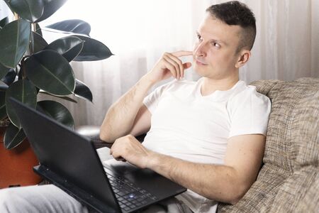 A focused European male freelancer takes notes studies working with a laptop, a young professional writes an essay in a notebook prepares for a test exam and sits at home on the couch. Remote workの写真素材