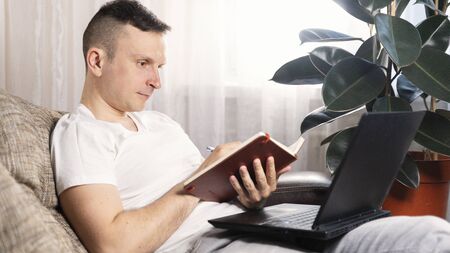 A focused European male freelancer takes notes studies working with a laptop, a young professional writes an essay in a notebook prepares for a test exam and sits at home on the couch. Remote workの写真素材