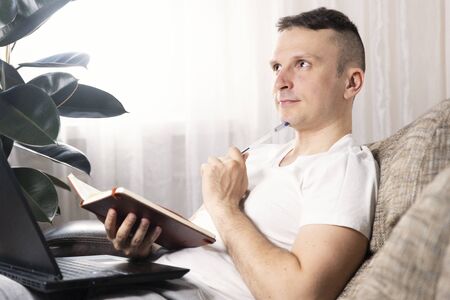 A focused European male freelancer takes notes studies working with a laptop, a young professional writes an essay in a notebook prepares for a test exam and sits at home on the couch. Remote workの写真素材