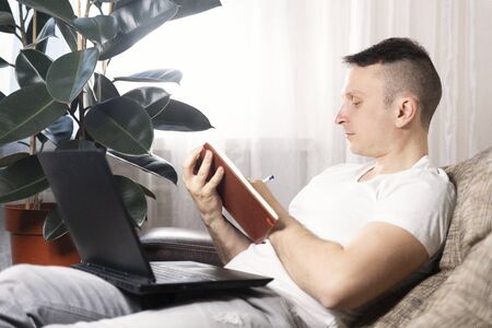 A focused European male freelancer takes notes studies working with a laptop, a young professional writes an essay in a notebook prepares for a test exam and sits at home on the couch. Remote workの写真素材
