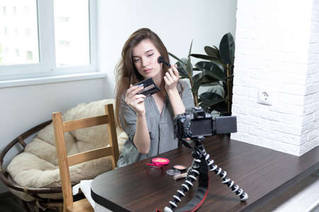 A beautiful girl is sitting in a bright room and making a selfie video for a fashion blog. The woman keeps a blog online and makes a photo. Fashion cosmetics reviewerの写真素材