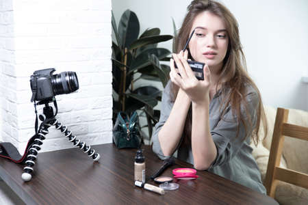 A beautiful girl is sitting in a bright room and making a selfie video for a fashion blog. The woman keeps a blog online and makes a photo. Fashion cosmetics reviewerの写真素材
