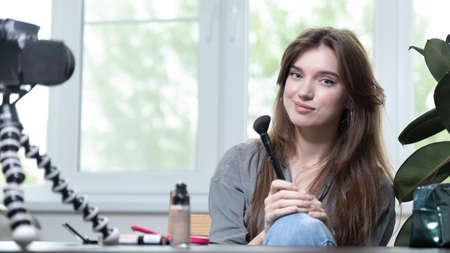 A beautiful girl is sitting in a bright room and making a selfie video for a fashion blog. The woman keeps a blog online and makes a photo. Fashion cosmetics reviewerの写真素材