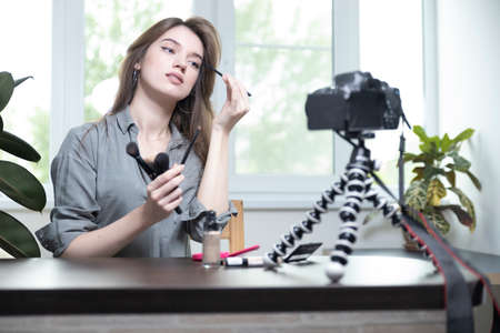A beautiful girl is sitting in a bright room and making a selfie video for a fashion blog. The woman keeps a blog online and makes a photo. Fashion cosmetics reviewerの写真素材