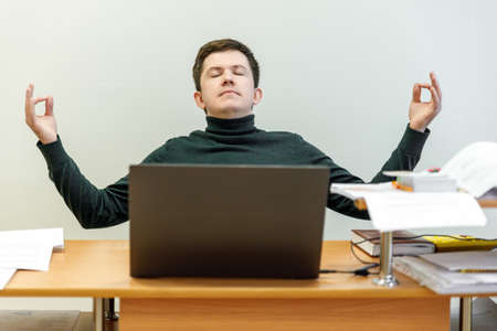 Relaxed corporate man sitting with closed eyes contemplating at work desk with laptop in office. Guy relaxing, relieves stress, gets positive thinking with yoga exercise. Stress free workplace conceptの写真素材