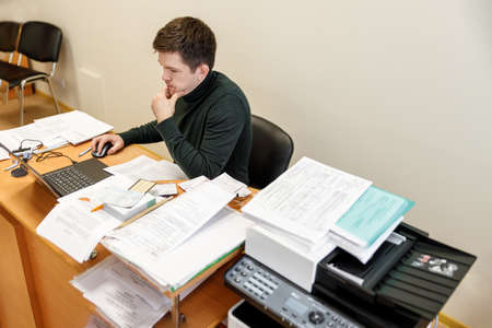 A casual businessman works in an office, sits at a Desk, types on a keyboard, looks at a computer screen. Concept of office work.の写真素材