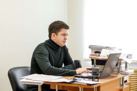 A casual businessman works in an office, sits at a Desk, types on a keyboard, looks at a computer screen. Concept of office work.の写真素材