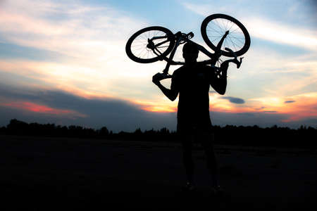 A silhouette of a sporty man standing in action lifting a Bicycle over his head in a meadow with the sun setting. Concept of sport and healthy lifestyleの写真素材