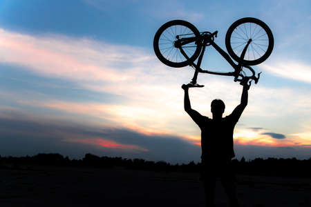 A silhouette of a sporty man standing in action lifting a Bicycle over his head in a meadow with the sun setting. Concept of sport and healthy lifestyleの写真素材
