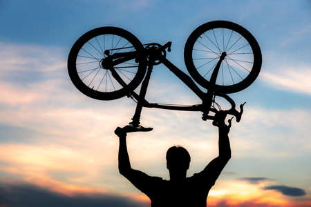 A silhouette of a sporty man standing in action lifting a Bicycle over his head in a meadow with the sun setting. Concept of sport and healthy lifestyleの写真素材