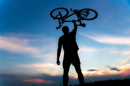 A silhouette of a sporty man standing in action lifting a Bicycle over his head in a meadow with the sun setting. Concept of sport and healthy lifestyleの写真素材