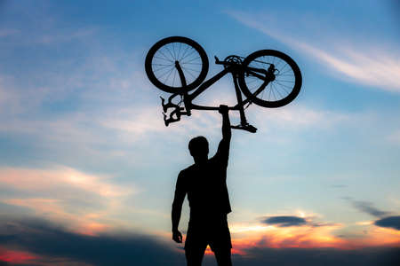 A silhouette of a sporty man standing in action lifting a Bicycle over his head in a meadow with the sun setting. Concept of sport and healthy lifestyleの写真素材
