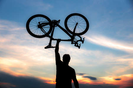 A silhouette of a sporty man standing in action lifting a Bicycle over his head in a meadow with the sun setting. Concept of sport and healthy lifestyleの写真素材