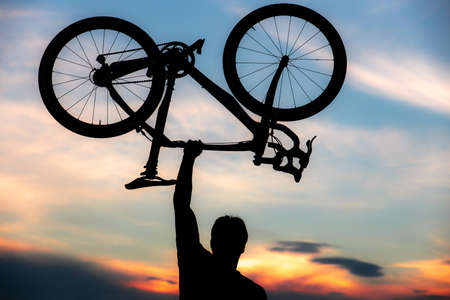 A silhouette of a sporty man standing in action lifting a Bicycle over his head in a meadow with the sun setting. Concept of sport and healthy lifestyleの写真素材