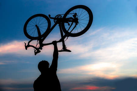 A silhouette of a sporty man standing in action lifting a Bicycle over his head in a meadow with the sun setting. Concept of sport and healthy lifestyleの写真素材
