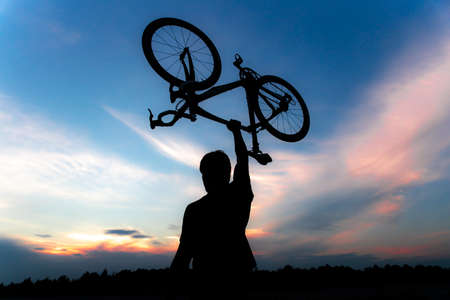 A silhouette of a sporty man standing in action lifting a Bicycle over his head in a meadow with the sun setting. Concept of sport and healthy lifestyleの写真素材