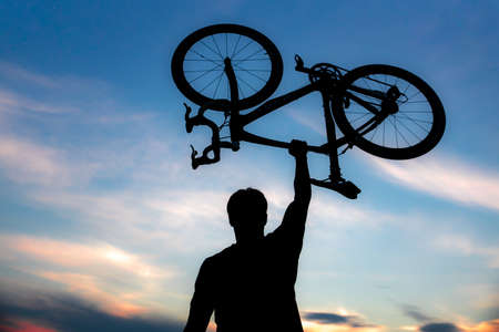 A silhouette of a sporty man standing in action lifting a Bicycle over his head in a meadow with the sun setting. Concept of sport and healthy lifestyleの写真素材