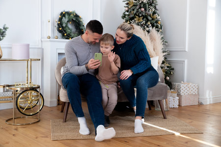 A cheerful family with their son laughs, watch videos together on the phone, a young dad holds a smartphone, loving parents have fun with the child. Video call, conversation with relativesの写真素材