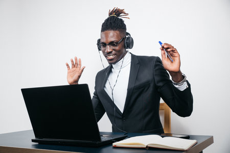 Smiling African American employee in headphones using laptop, looking at screen, making video call or watching webinar, writing notes, distance learning language concept, call center operator workingの写真素材