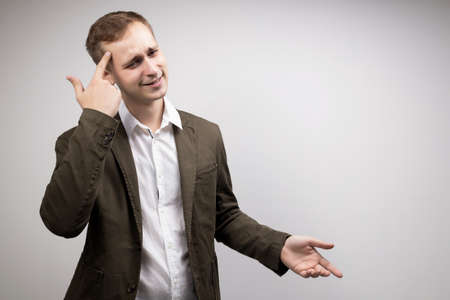 Young male businessman showing stupid gesture pointing finger to temple on gray studio background. Body language conceptの写真素材
