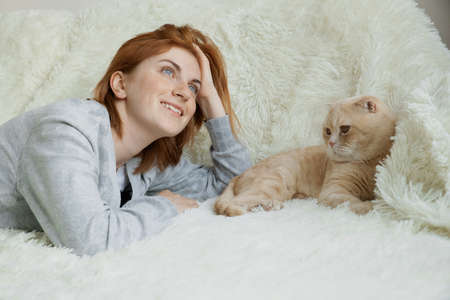 Happy dreamy young red-haired female lying on white comfortable plaid with ginger cat fluffy pet at homeの写真素材