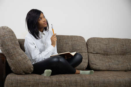 Thoughtful dreamy young african american female sitting on couch making notes, planning week to-do list write in notepadの写真素材
