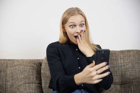 Happy excited young female looking at phone amazed by message about shopping or sale offer and good newsの写真素材