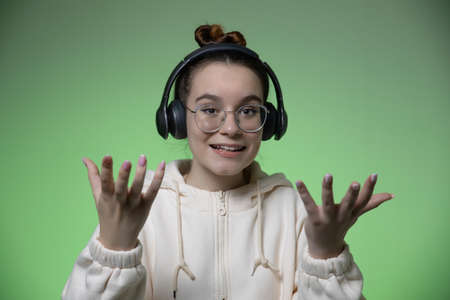 Bewildered young teen girl in headphones and glasses spread hands on green studio background with copy space for adの写真素材
