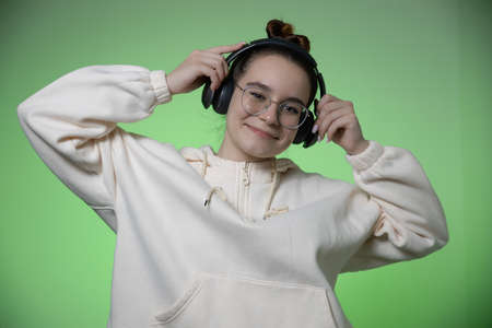 Smiling young teenage girl in modern wireless headphones having fun listen to music on green studio...