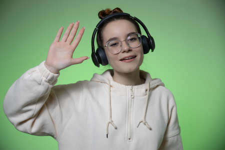 Smiling young teen girl blogger in glasses and modern wireless headphones on green studio background greeting wave handの写真素材