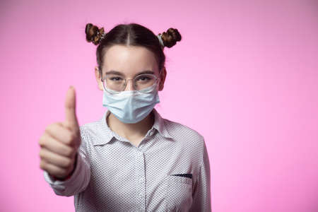 Stylish young teen girl recommending wear medical protective mask against coronavirus showing thumb up on pink backdropの写真素材